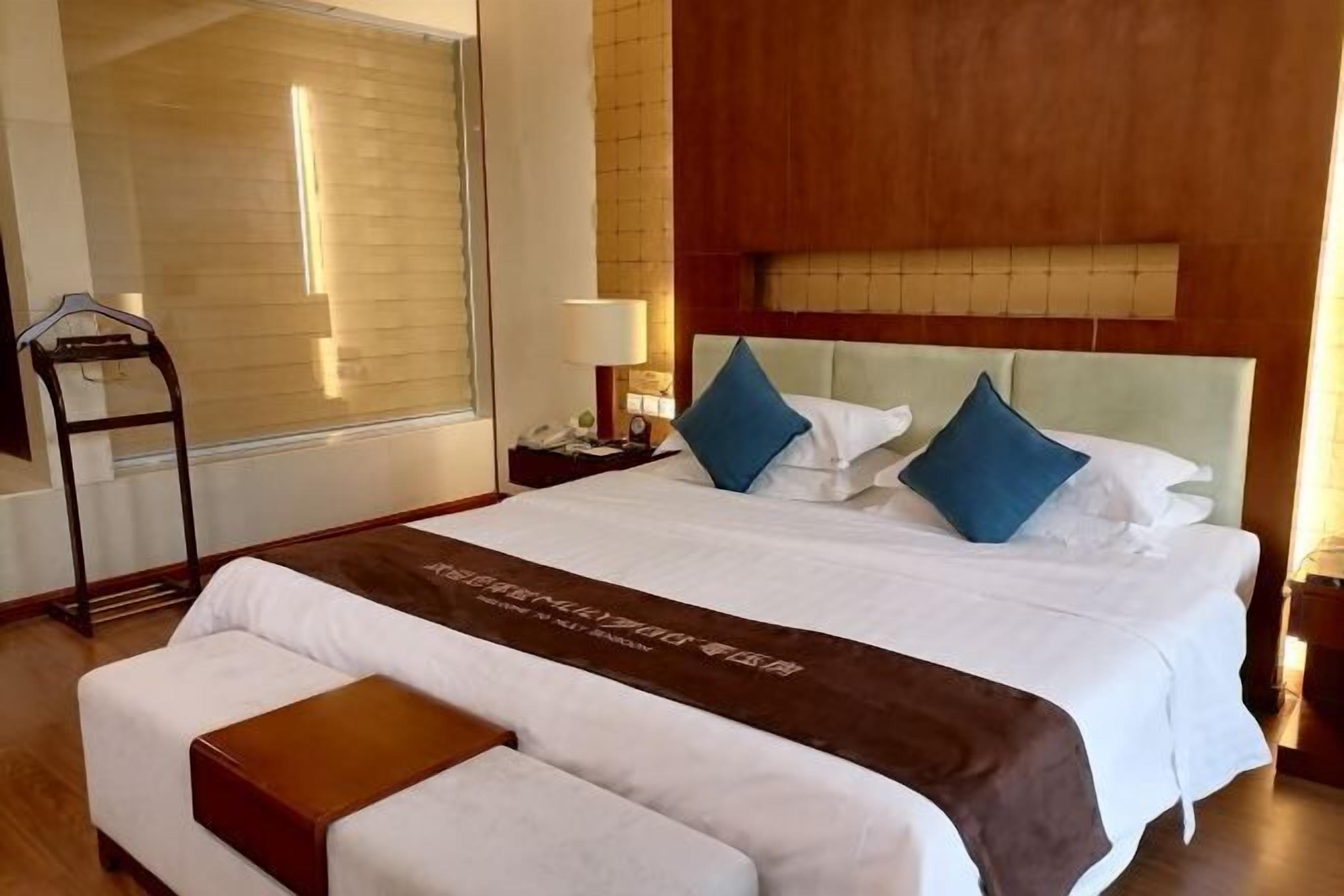 Une chambre d’hôtel avec un grand lit, une table de chevet avec une lampe, une tête de lit en bois et une fenêtre avec des stores.