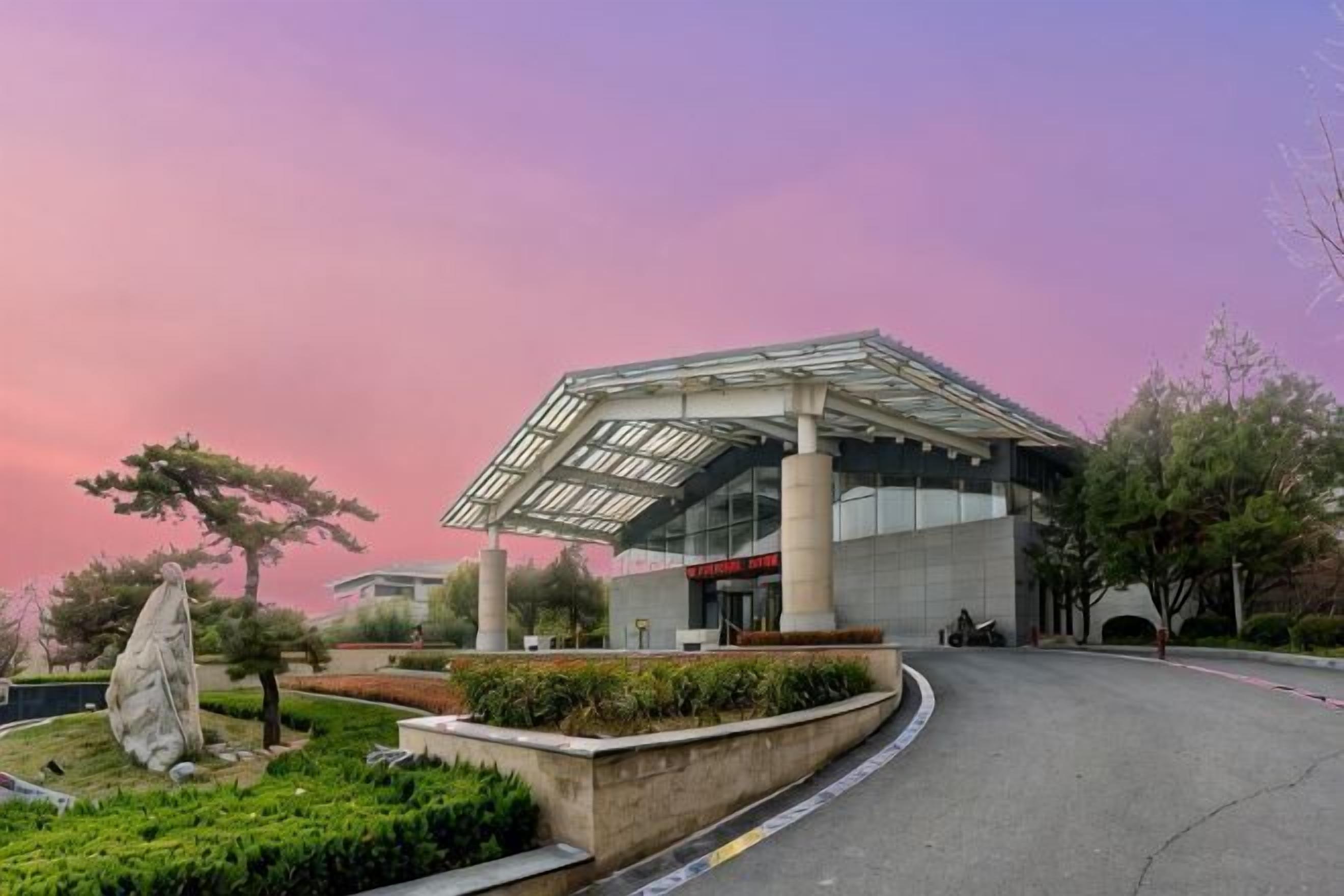 Un edificio moderno con techo de vidrio, rodeado de jardines ajardinados y un camino de entrada curvo.