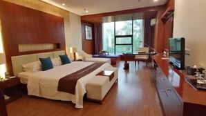 A hotel room with a large bed, a desk, a TV, and a window with a view of trees. - Bei Long Garden Hotel (Qingzhou)