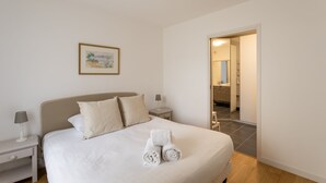 En pænt redt seng med hvide sengetæpper og sammenrullede håndklæder, et natbord med en lampe og et billede i ramme på væggen.