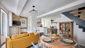 A modern living room with a yellow sofa, a round dining table set for four, and a kitchen area visible in the background.