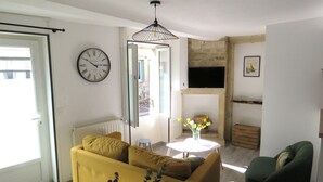 A living room with a yellow sofa, a round table with flowers, a clock, and a TV mounted on the wall.
