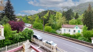 A road with cars, a white building with green shutters, and a forested hill in the background.