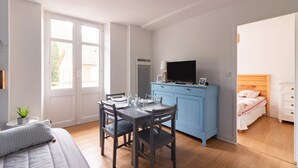 A room with a dining table, chairs, a TV on a blue cabinet, a bed with a wooden headboard, and a window with a view of a building outside.