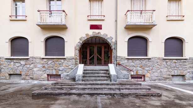 A building with a stone facade, a central entrance with a large arched doorway, and multiple balconies with railings.