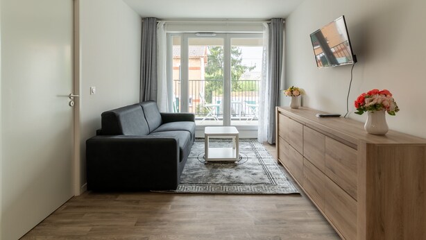 Un salon moderne avec un canapé noir, une petite table d’appoint blanche, un meuble en bois et un téléviseur fixé au mur.