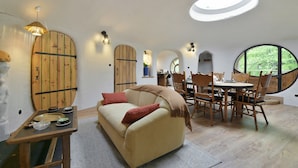 Ein gemütliches Interieur mit einer Couch, einem Esstisch und Stühlen, einem kleinen Holztisch mit Geschirr sowie einem großen Fenster mit Blick ins Grüne.