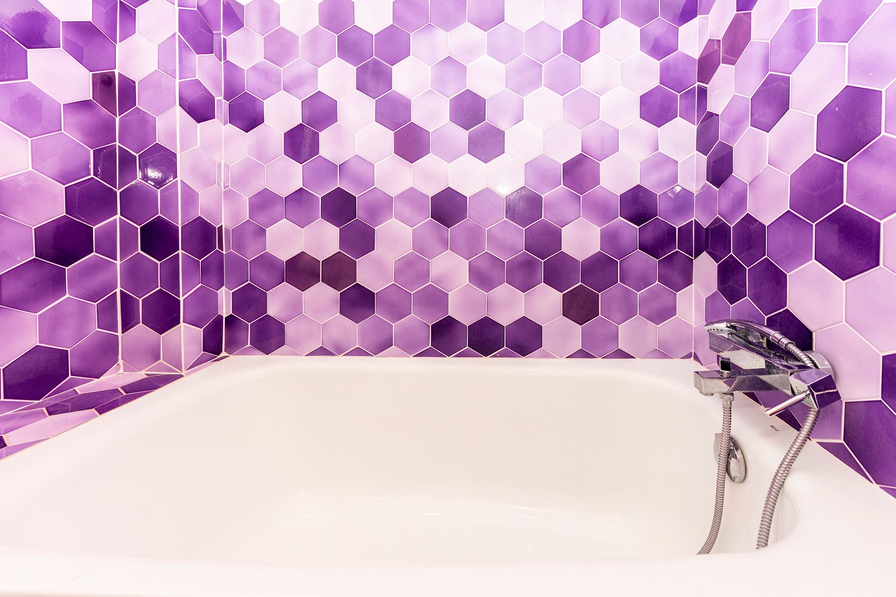 A bathroom with a white bathtub and a shower with a chrome faucet. The walls are tiled with purple hexagonal tiles.