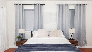 Un dormitorio con una cama, dos mesitas de noche con lámparas y cortinas grises.