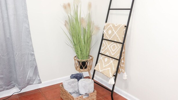 Esquina de una habitación con una cesta de mimbre que contiene velas, una planta en maceta y una escalera metálica negra con una toalla estampada.