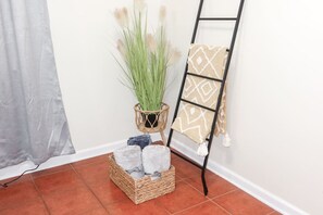 A corner of a room with a wicker basket containing candles, a potted plant, and a black metal ladder with a patterned towel. - 1Bed 1Bath Apt near EWR, Parking 18' from Penn St. (Newark)