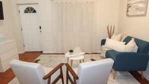 Una sala de estar con un sofá azul, sillas blancas, una mesita pequeña y una puerta blanca con ventanal.