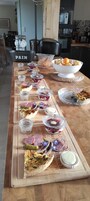 Uma longa mesa de madeira com diversos alimentos e acompanhamentos, incluindo uma tigela de frutas, uma torta e vários pequenos potes de compotas.