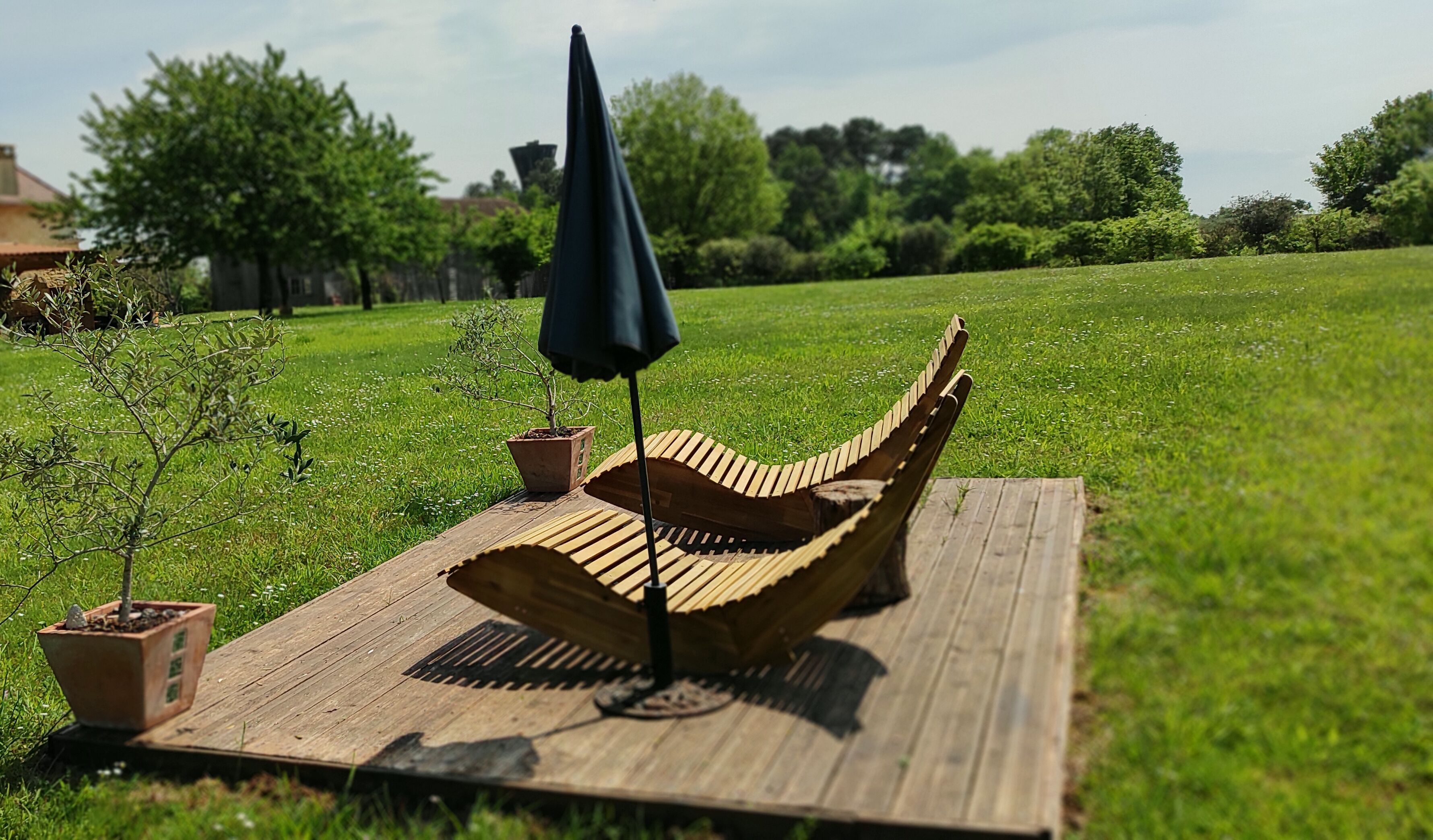 Eine Hängematte auf einer Holzterrasse inmitten von Wiese und Topfpflanzen.