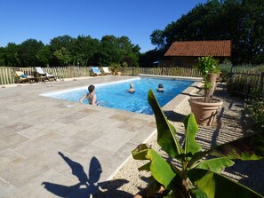 A swimming pool with tiled flooring, surrounded by a wooden fence, potted plants, and a building with a tiled roof in the background. - BRITAVIT Chambres et Table d'Hôtes (Saint-Avit-Senieur)