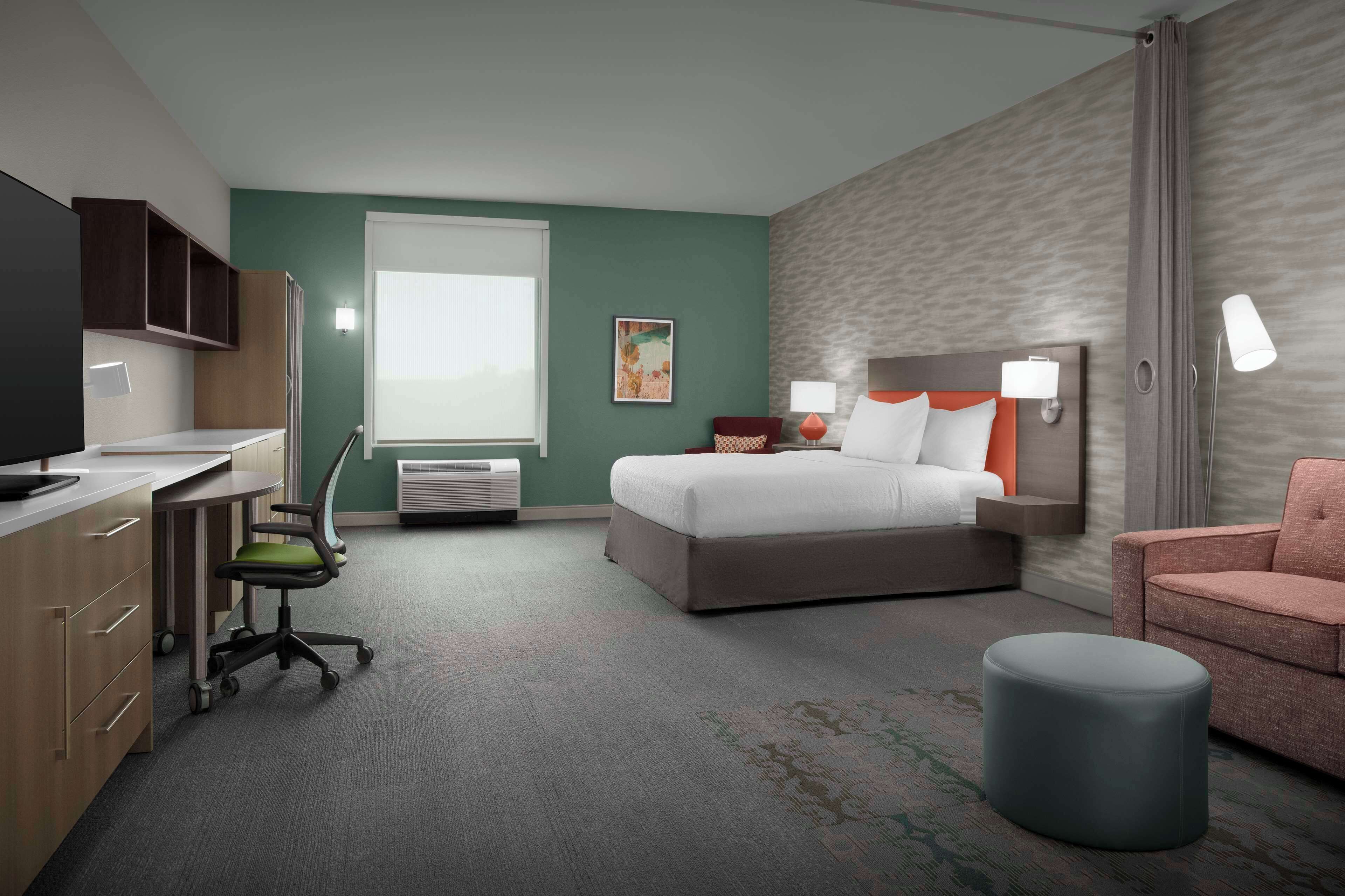 A hotel room with a bed, a desk, a chair, a small ottoman, and a window.