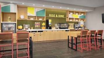 A modern kitchen with a 'RISE & DINE' sign, wooden cabinets, a coffee machine, and red chairs.