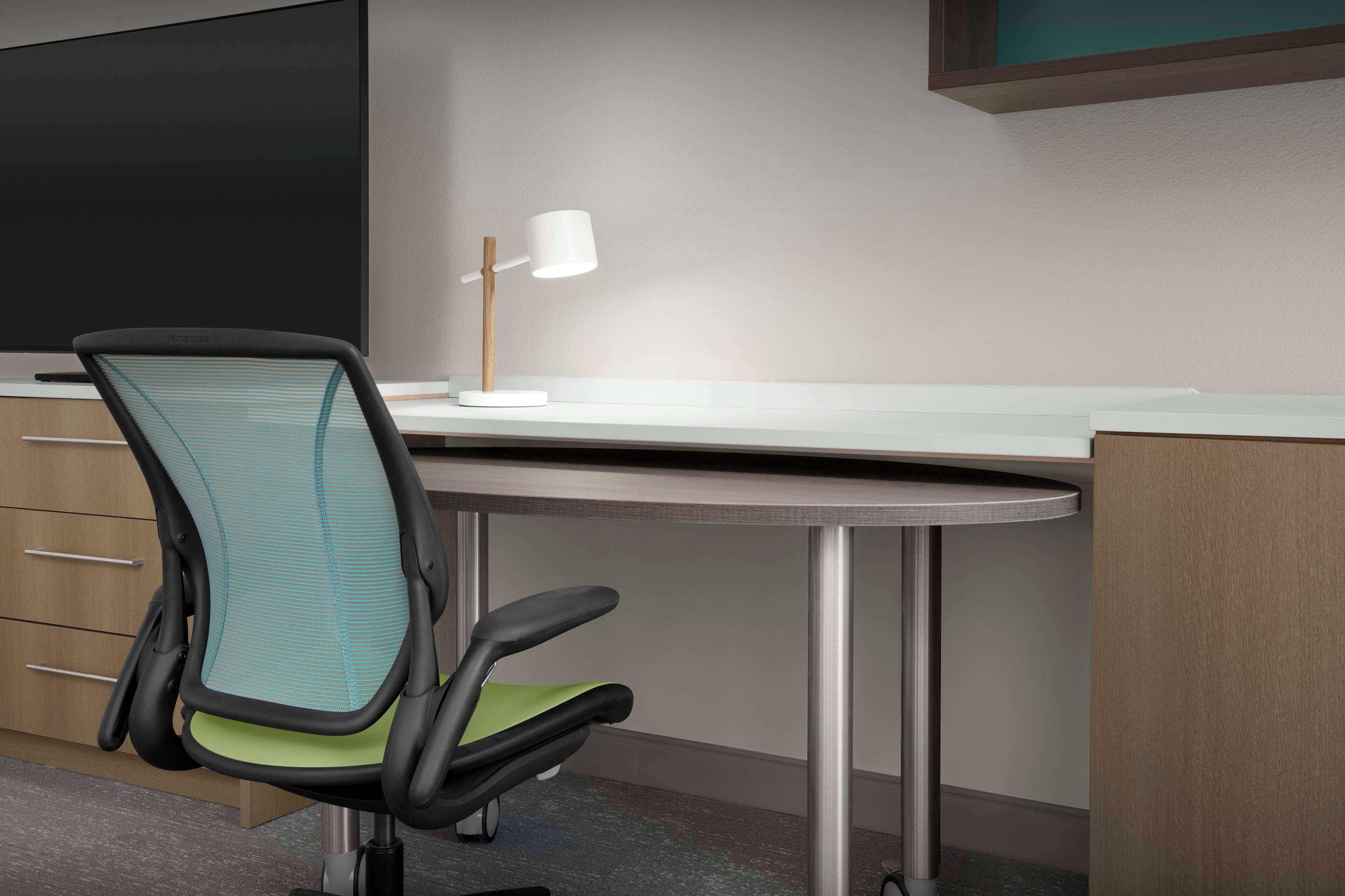 A modern office chair with a mesh backrest and green seat cushion, a white desk lamp, and a flat-screen monitor on a white desk.