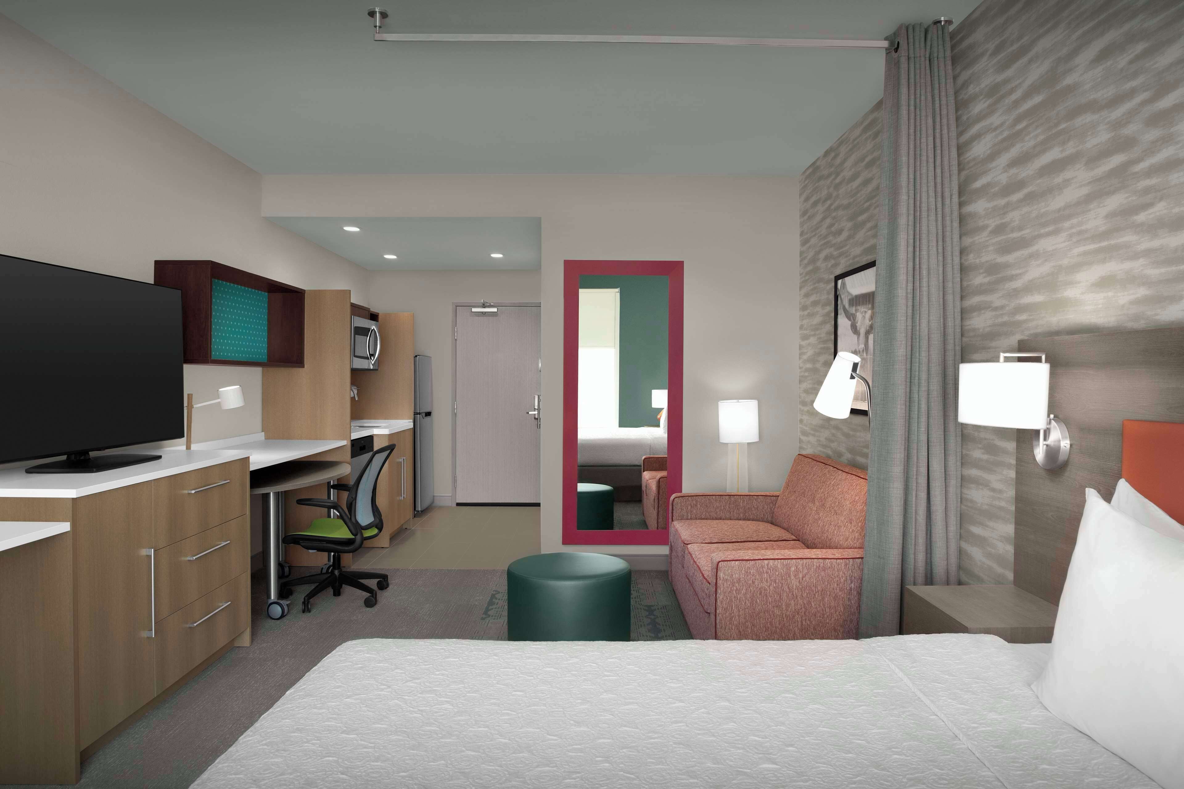 A hotel room with a bed, a desk with a computer, a TV, a sofa, a mirror, and a nightstand with a lamp.