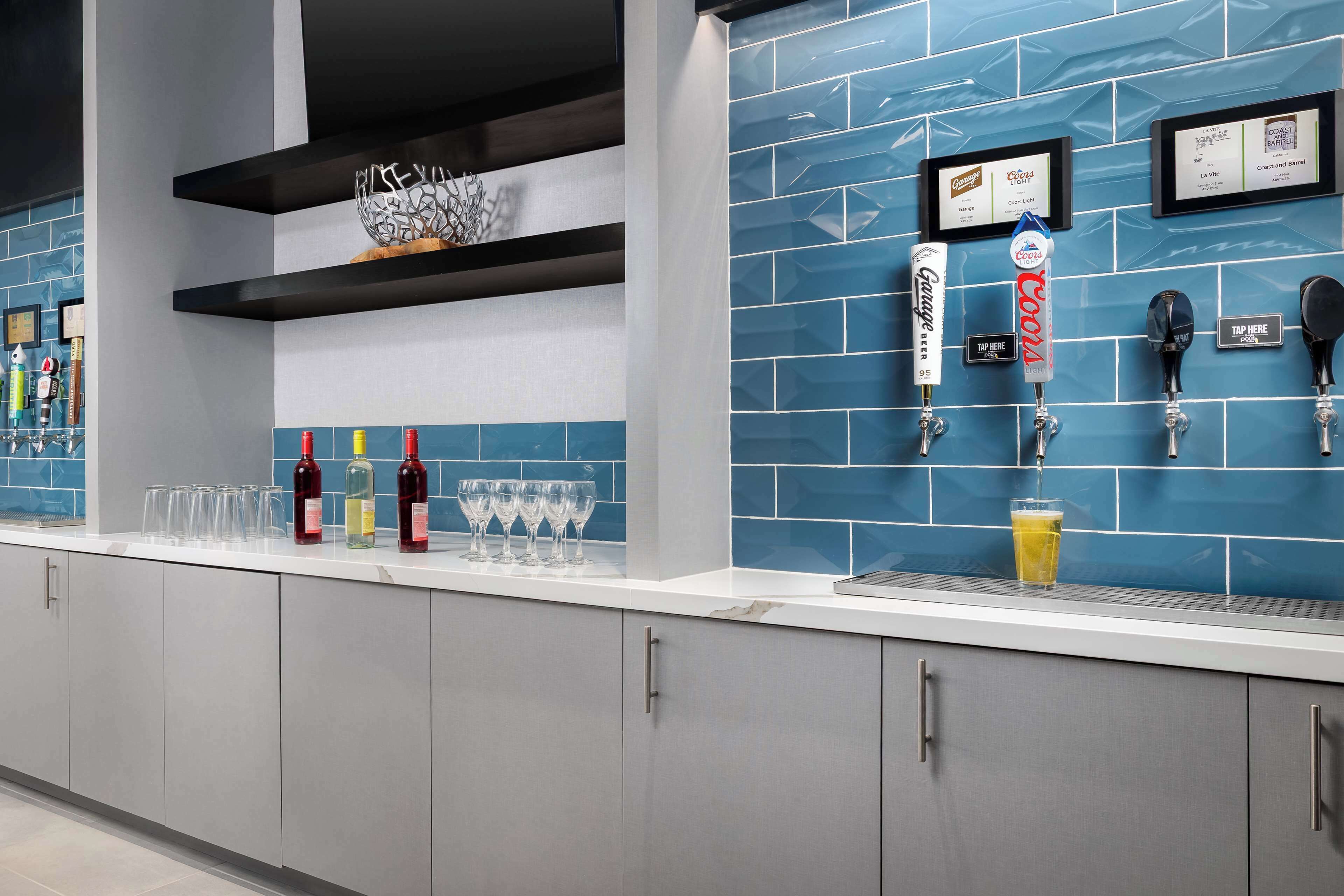 Una zona de bar con azulejos azules, una barra con vasos y botellas, y una grifo de cerveza Coors.