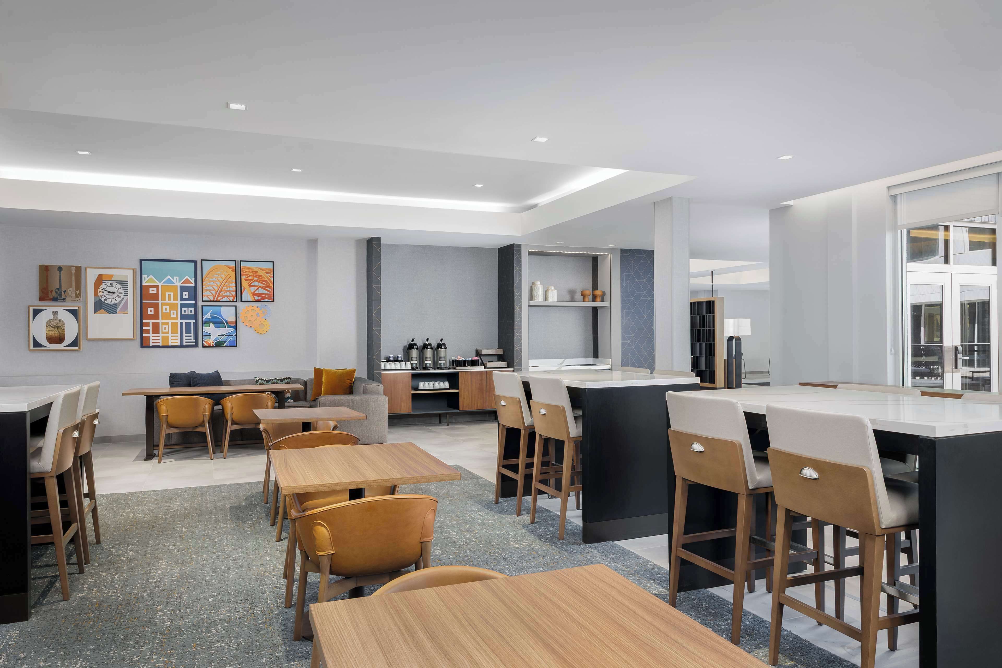 A modern dining area with wooden tables and chairs, a kitchen in the background, and artwork on the walls.