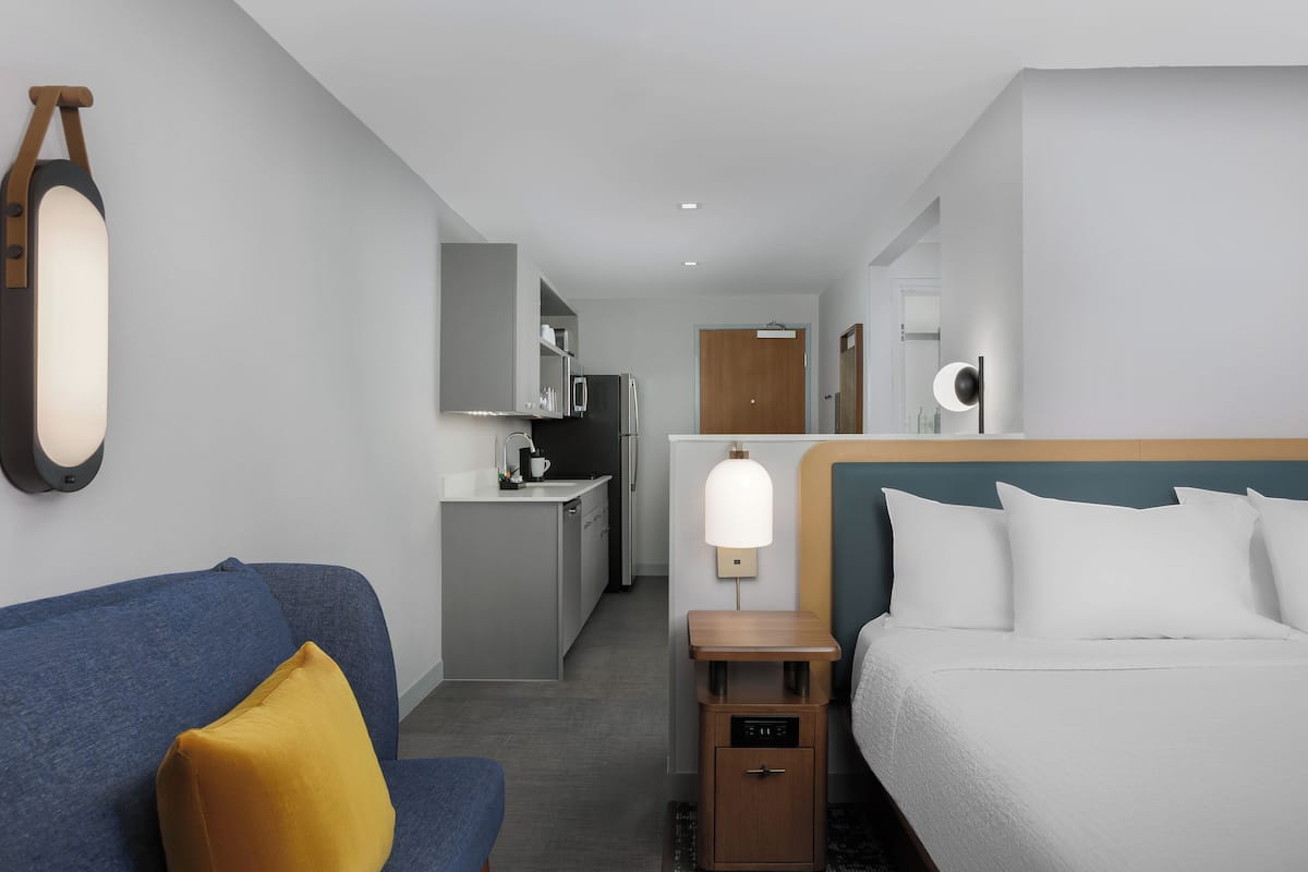 Una habitación de hotel moderna con un sofá azul, una cama con ropa de cama blanca, una mesita de noche de madera y una kitchenette al fondo.