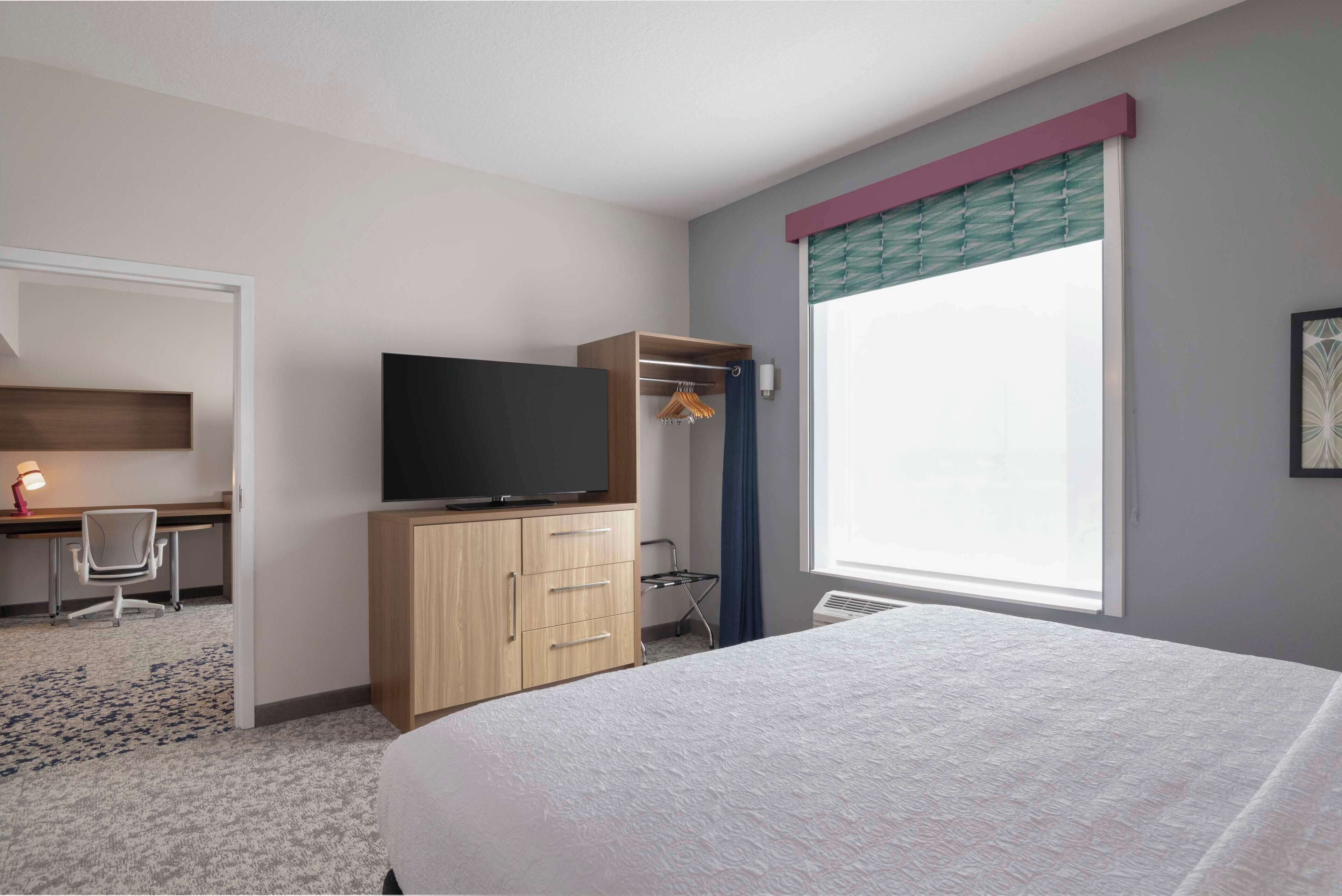 A hotel room with a bed, a desk with a chair, a television, a wardrobe, and a window with curtains.