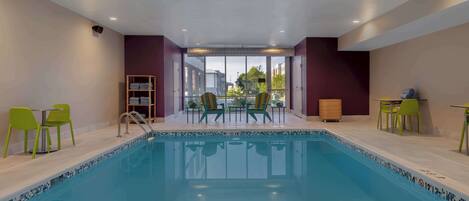 A swimming pool with chairs and tables, a wooden cabinet, and a view of buildings through large windows.
