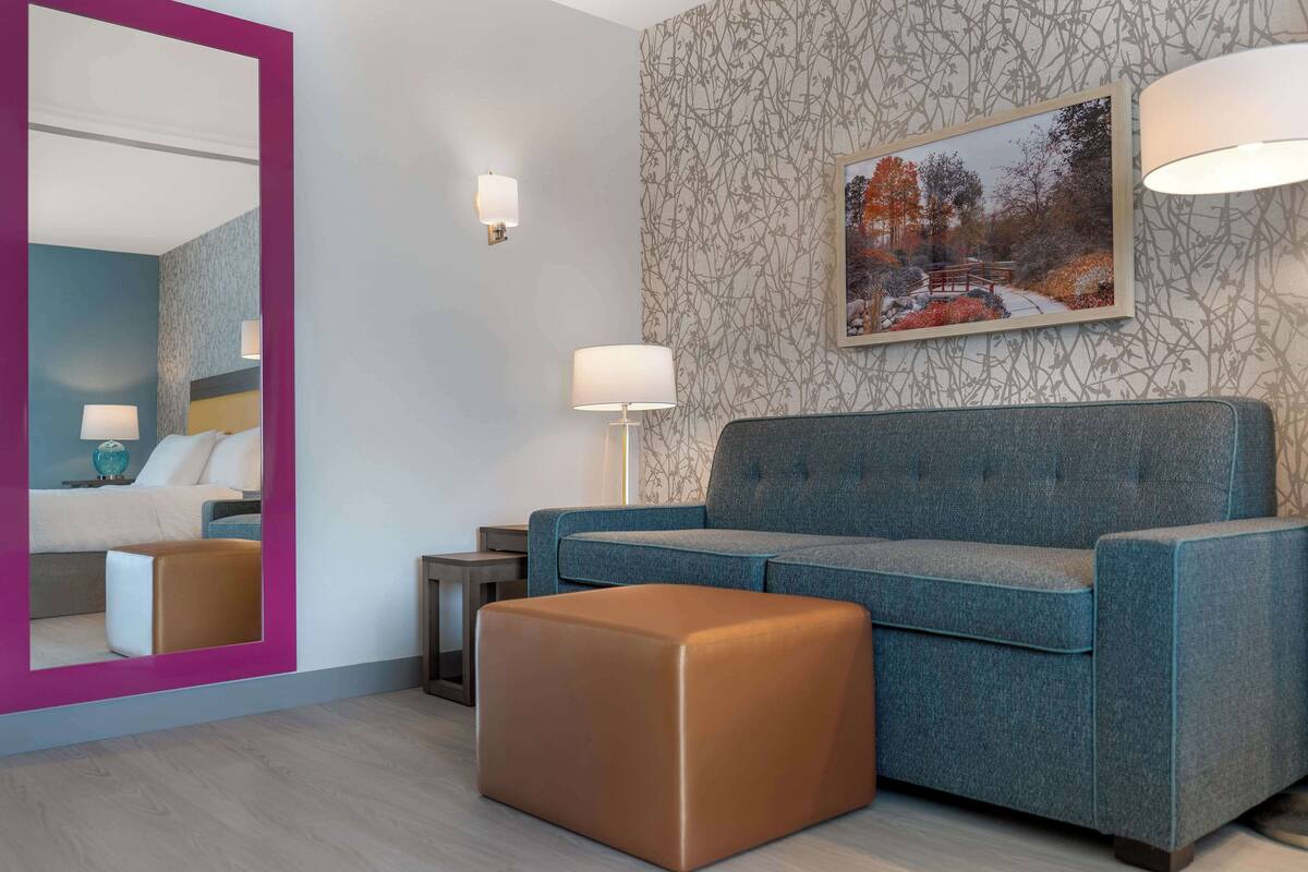 Ein modernes Hotelzimmer mit einem großen Spiegel, einem blauen Sofa, einem braunen Hocker und einem gerahmten Bild an der Wand.