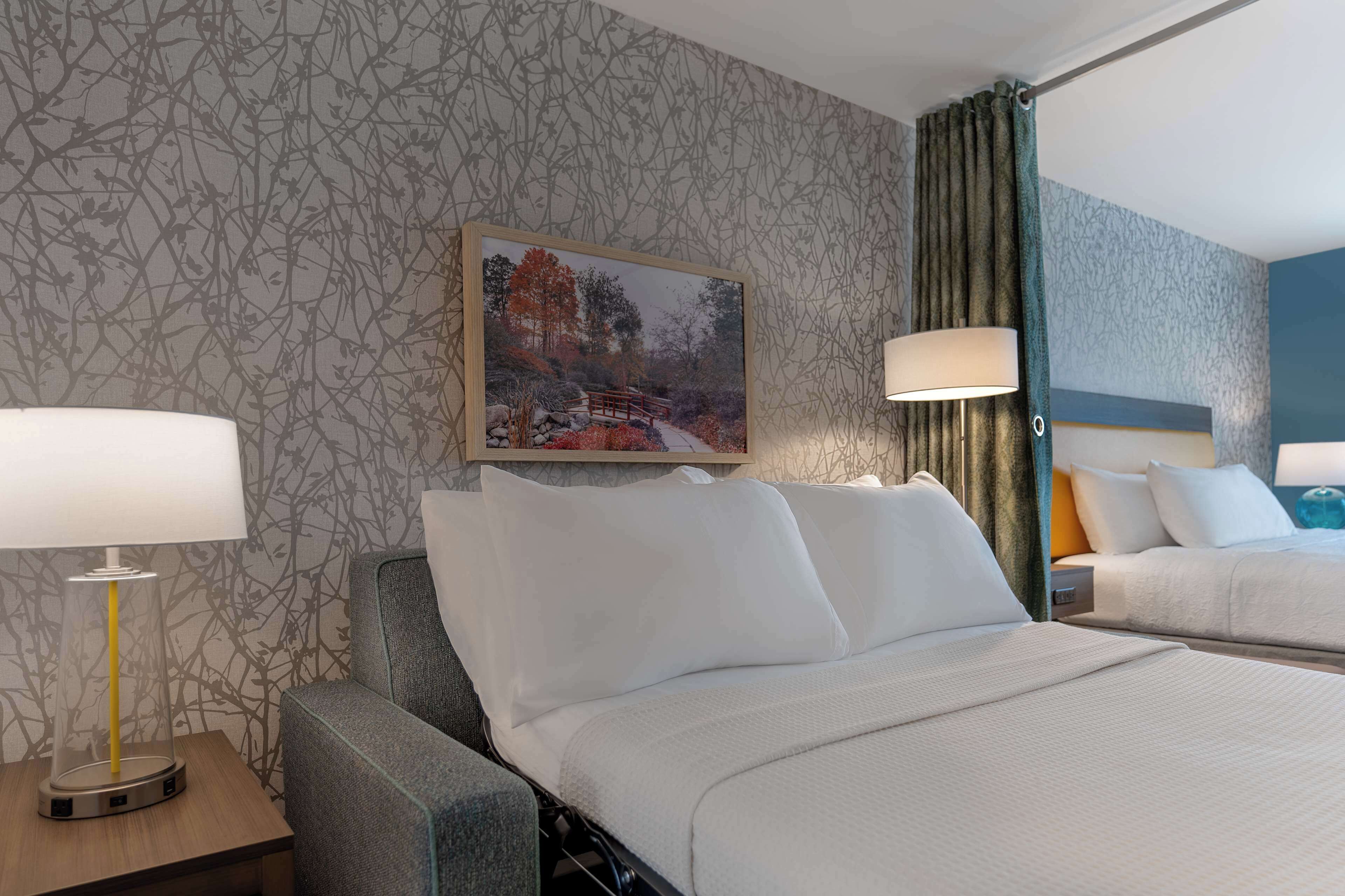 A hotel room with a bed, a chair, a nightstand, a lamp, and a painting on the wall.