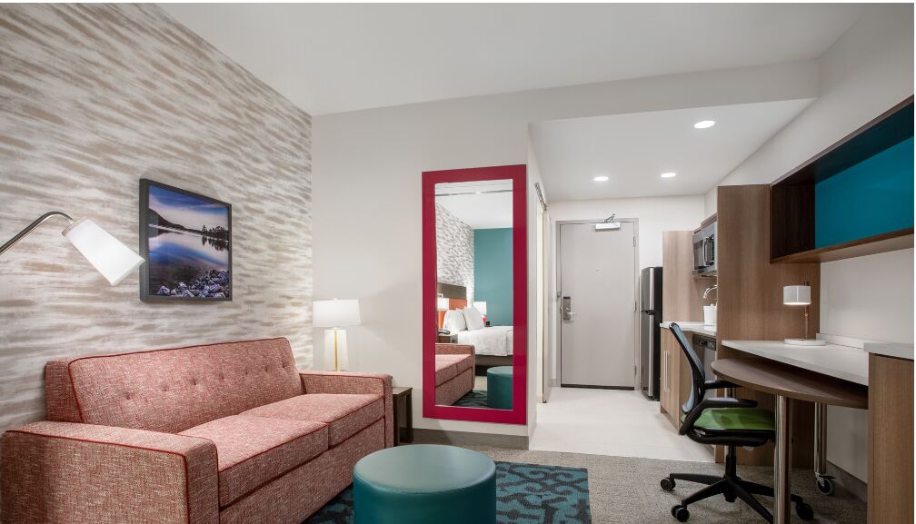 A modern hotel room with a sofa, a desk, a mirror, and a kitchenette.