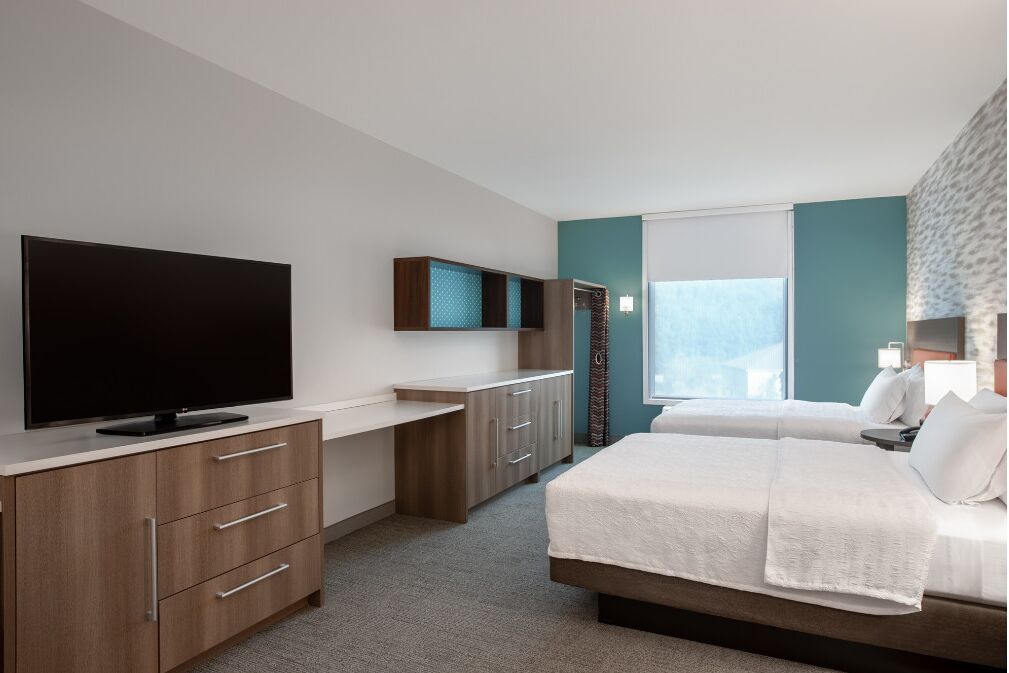 A modern hotel room with a flat-screen TV, a desk with drawers, a bed with white bedding, and a window with a view.