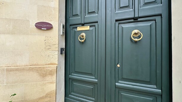 Two dark green doors with brass knockers, a plaque above the left door, and a small plant on the ground.