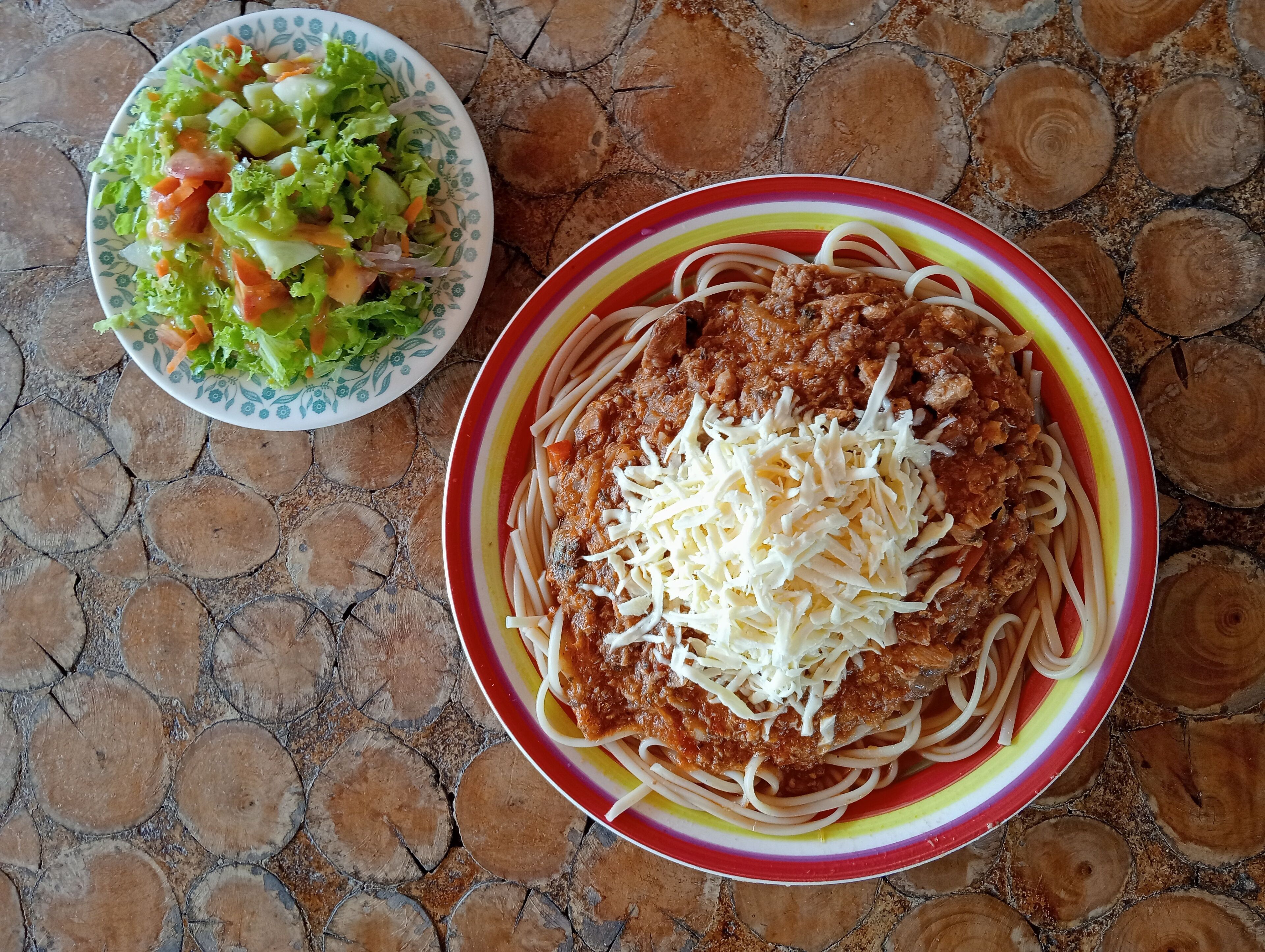 A plate of spaghetti with meat sauce and shredded cheese, and a side salad on a stone-tiled table.