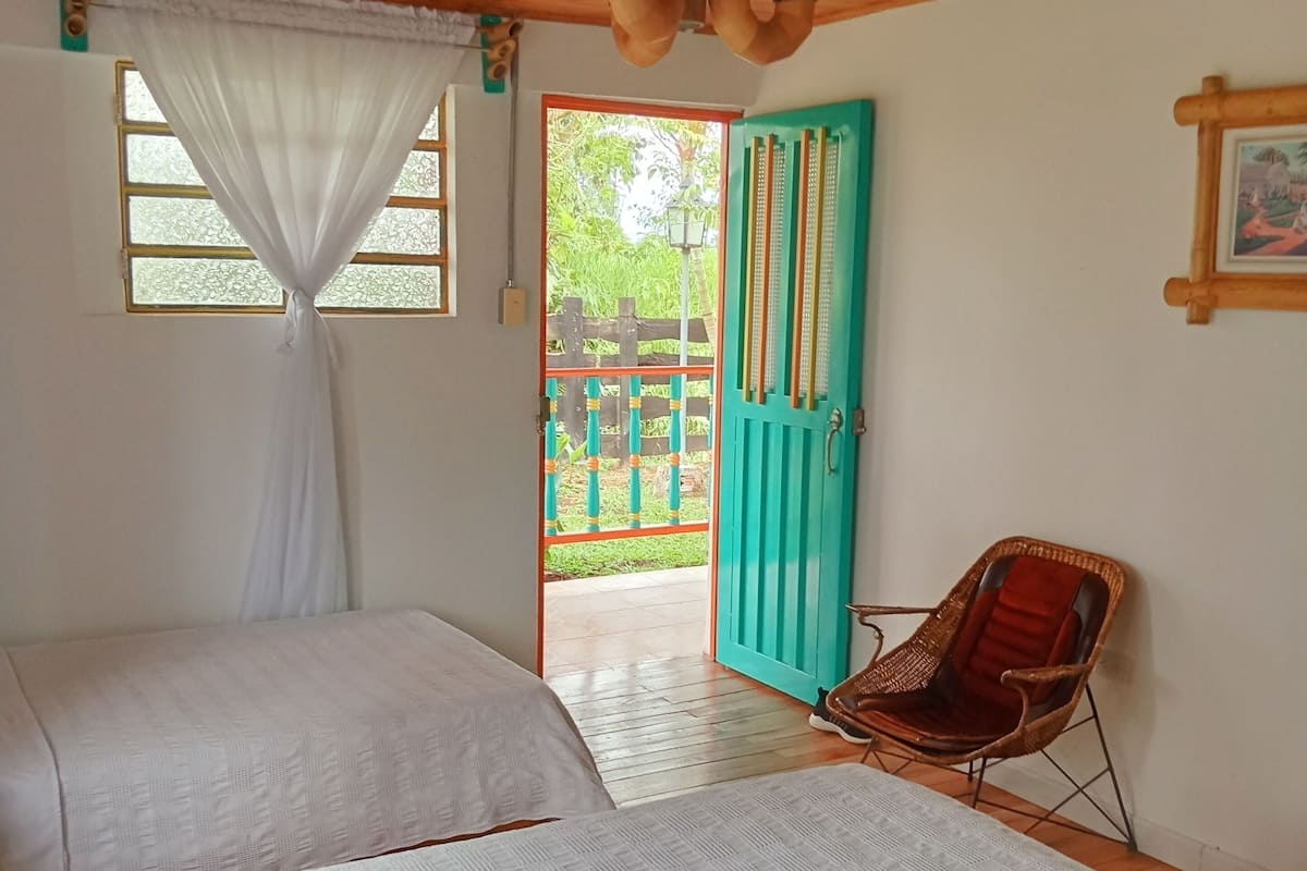 Une pièce avec deux lits, un plafond en bois, une fenêtre avec des rideaux, une porte donnant sur l’extérieur et une chaise en osier.