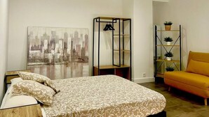 Un dormitorio con cama, un cuadro de paisaje urbano, un perchero negro, un sofá amarillo y una estantería con plantas.