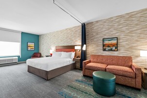 Habitación de hotel con cama, sofá, reposapiés redondo, mesita de noche, lámpara y un cuadro en la pared.