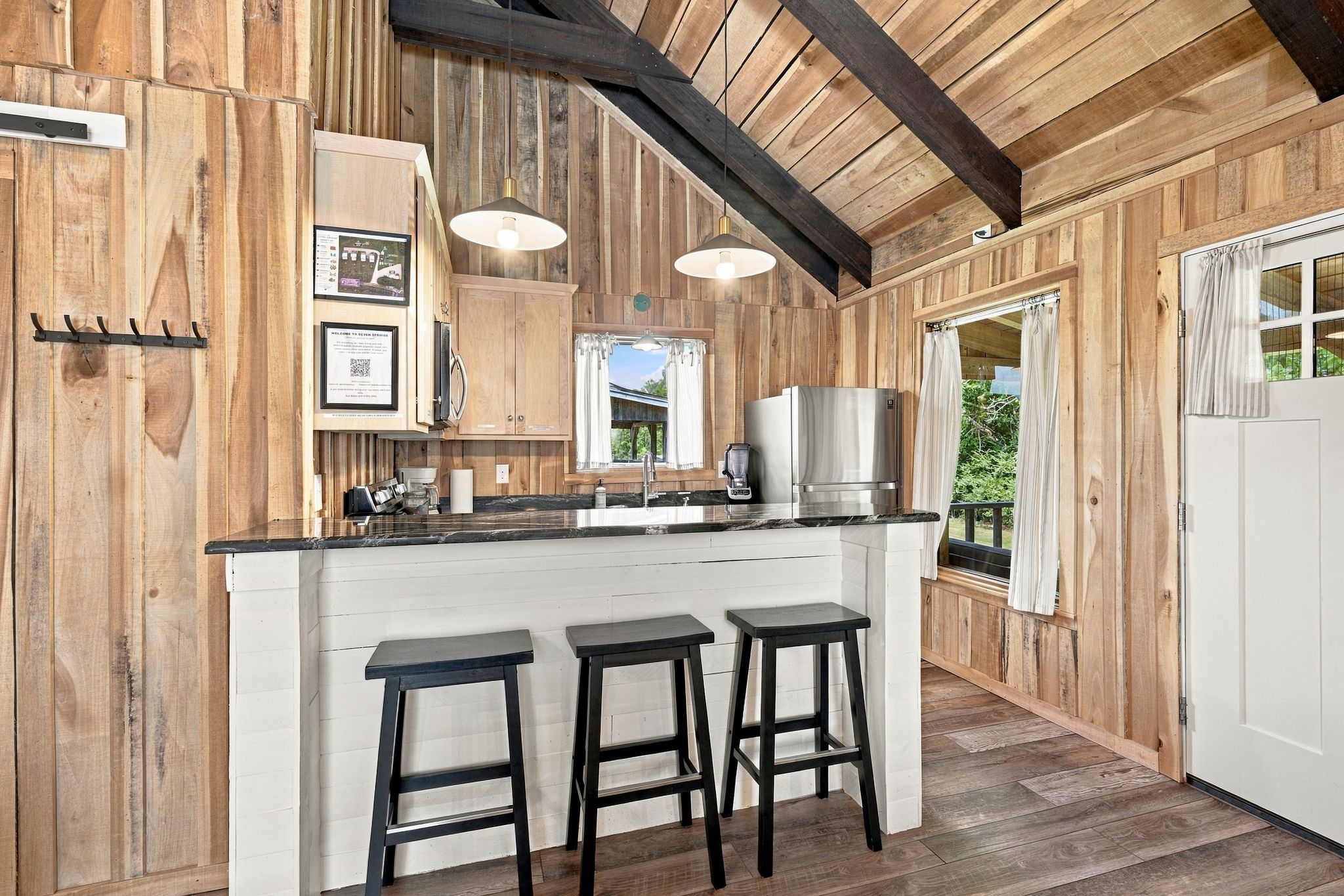 Un intérieur en bois avec un coin cuisine comprenant un bar avec des tabourets, un réfrigérateur et une fenêtre avec des rideaux.