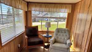 A cozy room with wooden walls, a window with blinds, a cushioned armchair, a small table with a lamp, and a view of a backyard with a vehicle. - Rocky Racoon's Rustic Retreat at Sage Lake!  Dock slip & tons of fun await!! (Hale)