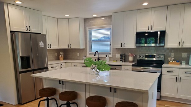 A modern kitchen with white cabinets, stainless steel appliances, a central island with stools, and a window with blinds. - Cozy 3 BR in center of town w/work space. Walking distance to main attractions  (Peachtree City)