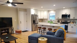 A modern kitchen with white cabinets, a stainless steel refrigerator, a microwave, and a dishwasher. There is a blue sofa with a yellow throw blanket, a small round ottoman, and a black coffee table. - Cozy 3 BR in center of town w/work space. Walking distance to main attractions  (Peachtree City)