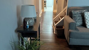 En hall med ett mörkt trägolv, en lampa, en krukväxt och en grå soffa med mönstrade kuddar.