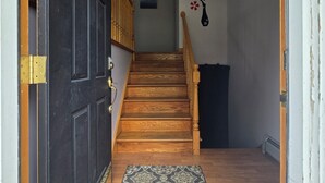 Un escalier en bois avec une rampe, une porte noire dotée de charnières en laiton, et un tapis à motifs menant à une pièce dont le mur est blanc et qui est décorée d’un motif floral.