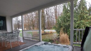 Un bain à remous sur une terrasse en bois, avec un patio couvert, entouré d’arbres et d’une clôture blanche.