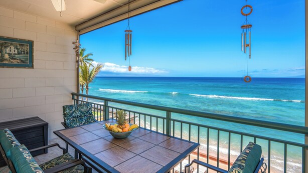 A balcony with a wooden table and chairs, a bowl of fruit, and a view of the ocean.