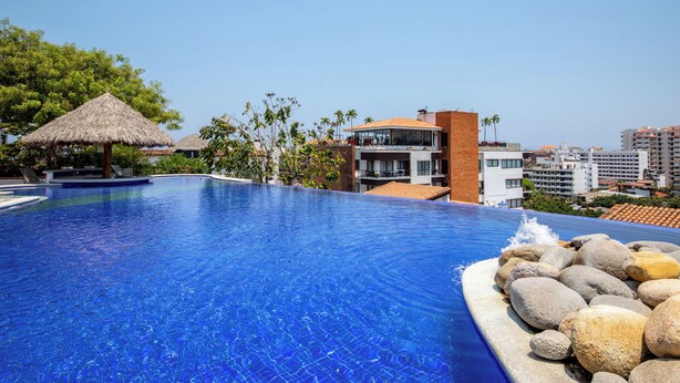Una piscina con superficie de agua azul cristalina, una sombrilla de paja y vista a edificios modernos al fondo.