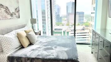 A modern bedroom with a large bed, a dresser, and a city view through large windows.