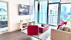 Ein modernes Wohnzimmer mit einem großen Fernseher, einem roten Sessel, einem pinkfarbenen Hocker und einem beigen Sofa.