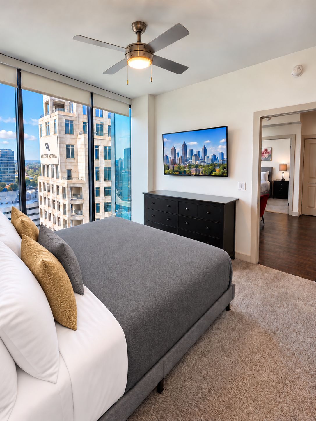 Une chambre avec un grand lit, un ventilateur de plafond, un tableau représentant un paysage urbain et une vue sur un immeuble de grande hauteur.