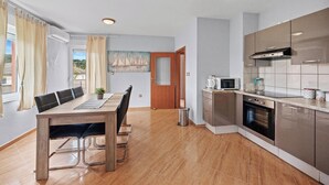 A modern kitchen with stainless steel appliances, a dining area with a wooden table and chairs, and a view of the outside through a window with curtains. - Seafront Apt, Tastefully Furnished - Neda Smilje (Kampor)